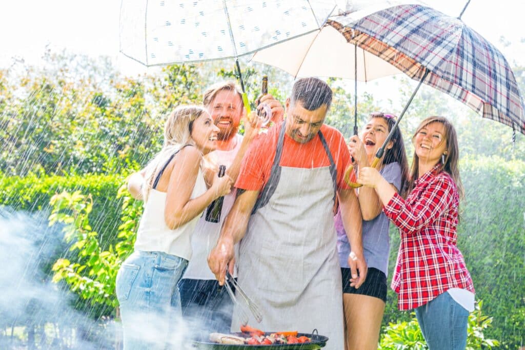 Eine Gruppe von Freunden grillt im strömenden Regen, geschützt durch Regenschirme, wobei ein Pavillon wasserdicht den idealen Schutz bieten würde.