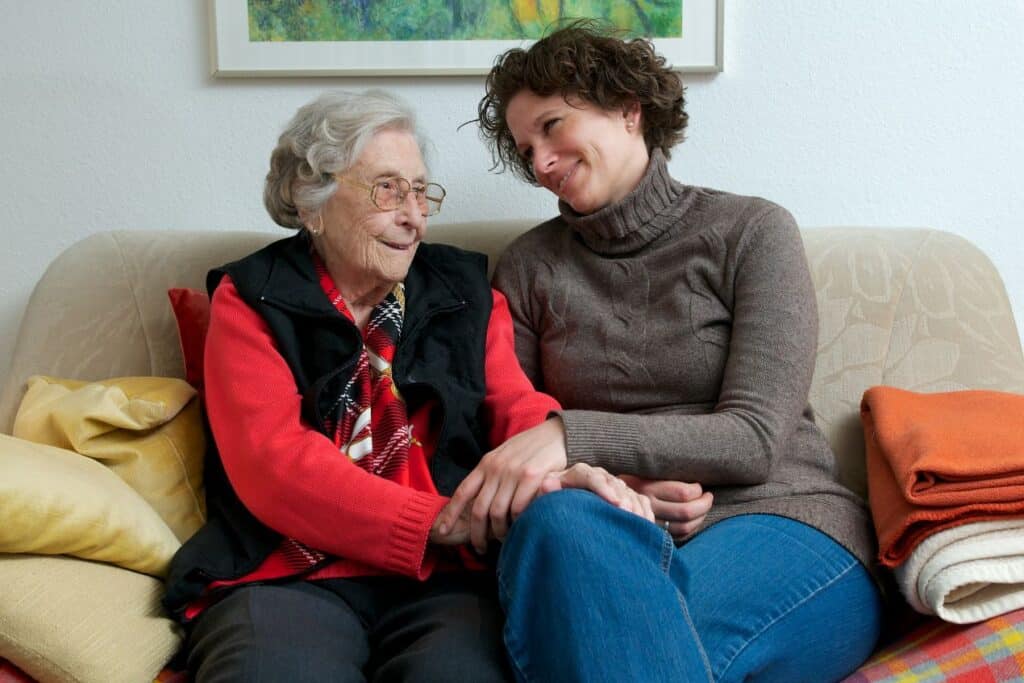 Ältere Frau und Tochter sitzen auf dem Sofa und halten sich an den Händen – Symbolbild für liebevolle häusliche Betreuung mit 24 Stunden Pflege Frankfurt